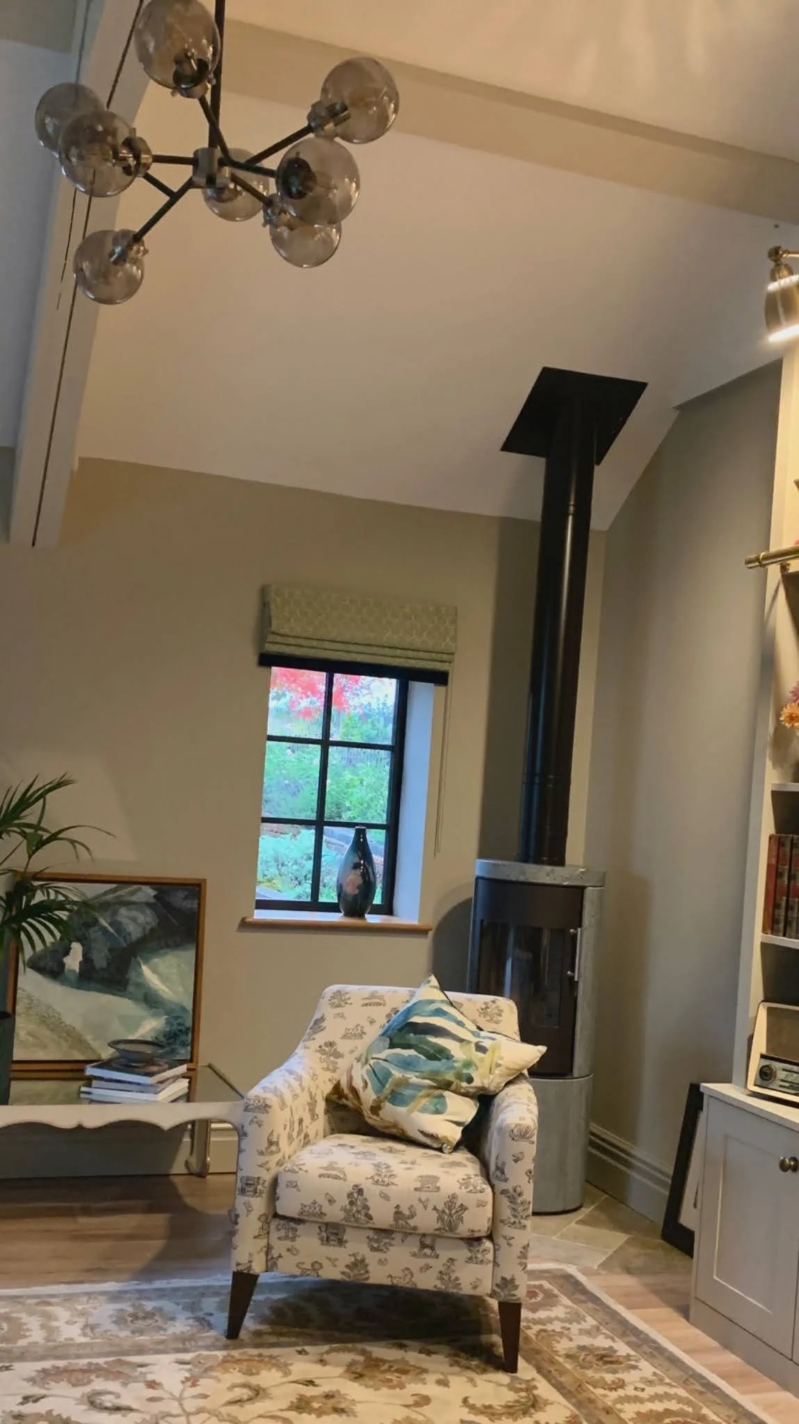 A vaulted sitting room with a sculptural glass-globe chandelier, patterned armchair and cushions beside a log burner, and an arched window dressed with a botanical roman blind