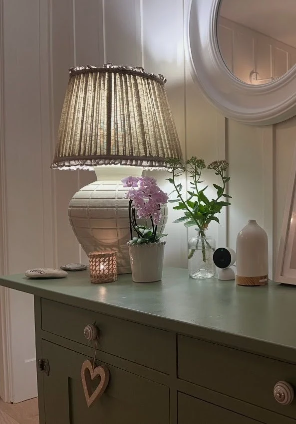 A pleated lampshade glowing on a soft green painted table beside an orchid, cut stems and a flickering candle — small details layered together