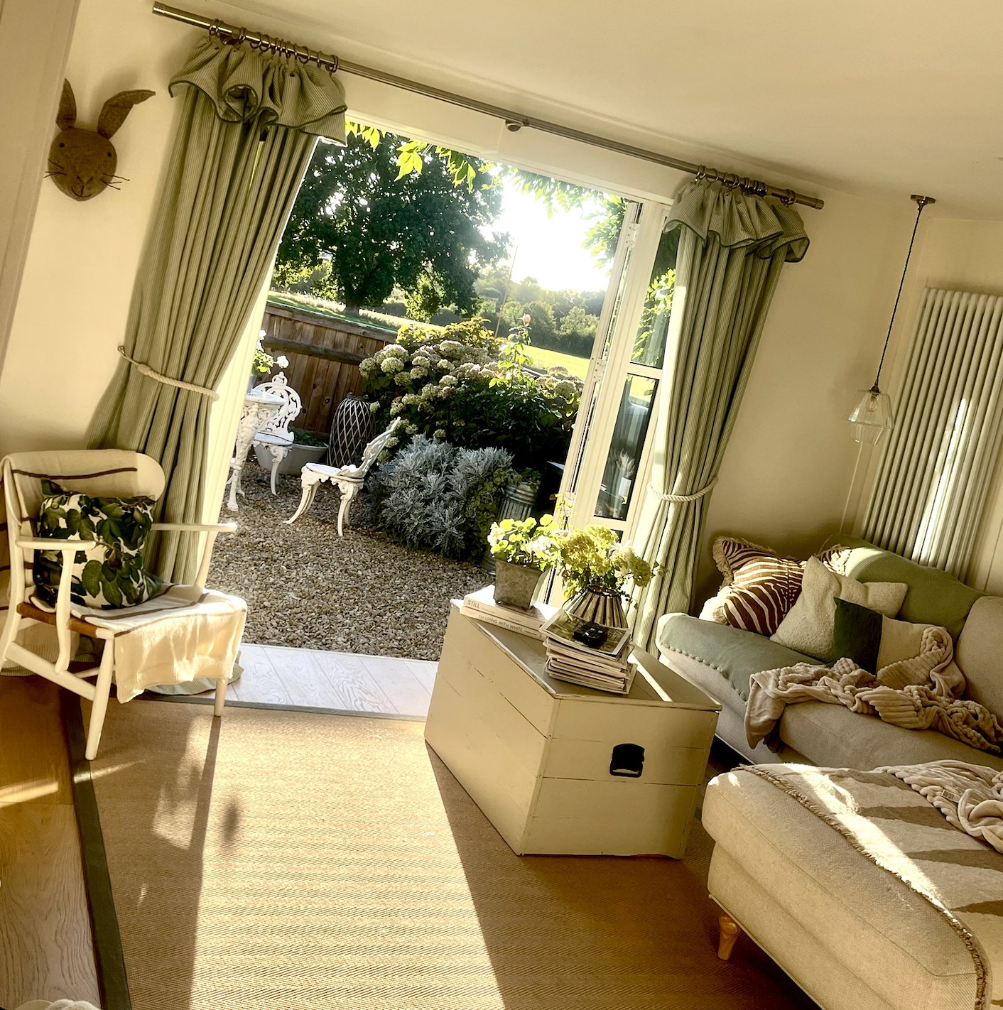 Sunlit sitting room with layered cushions, throws, curtains, and open French doors to the garden