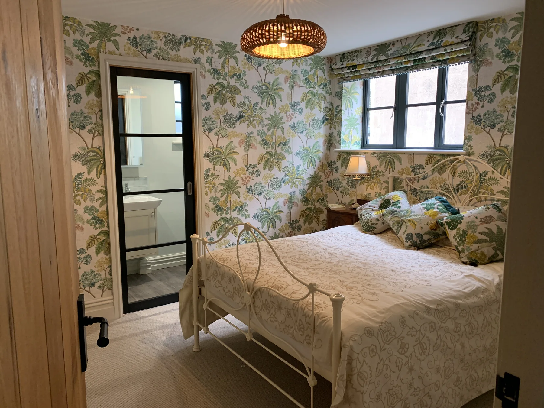 Botanical guest room with palm leaf wallpaper, white iron bed, and ensuite beyond