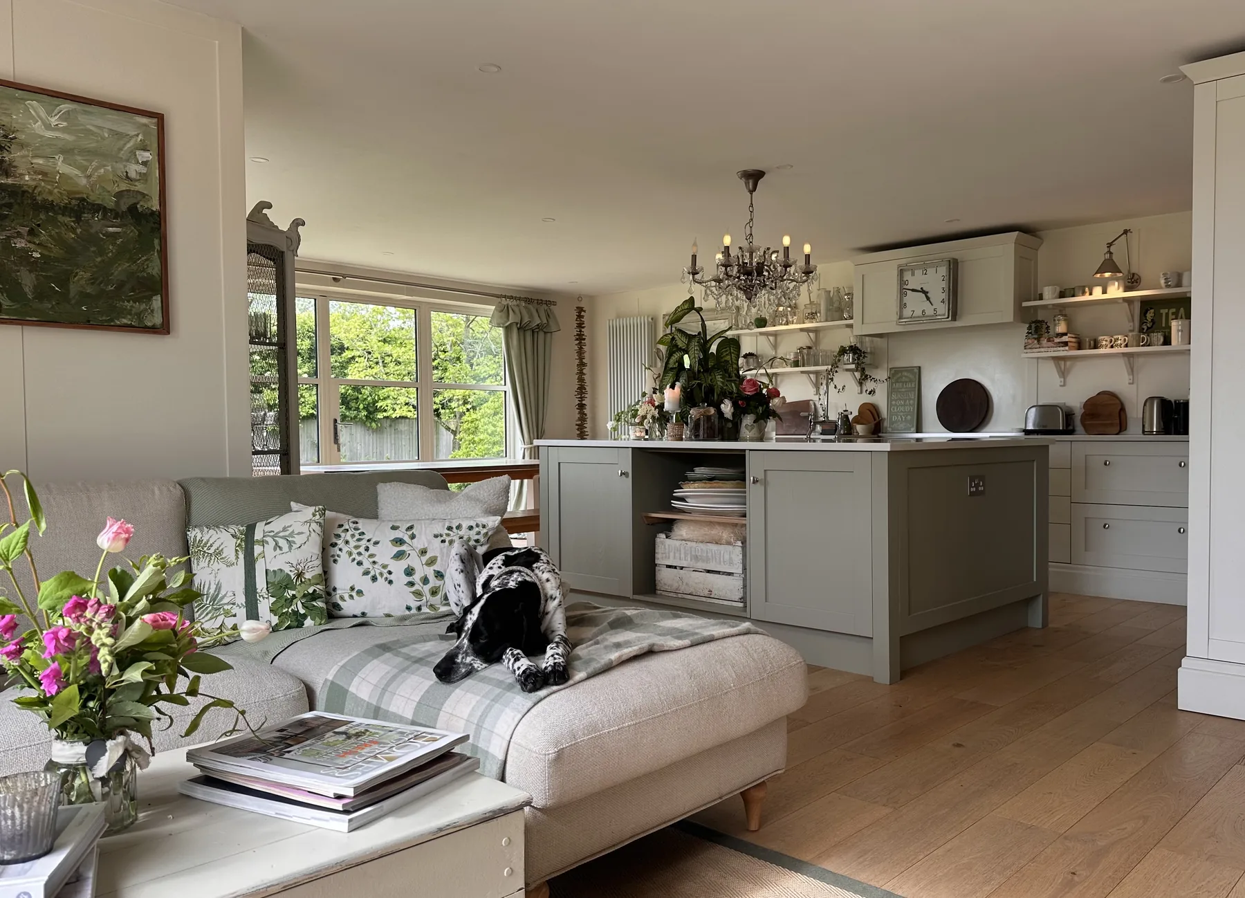 Appletreewick — open-plan sitting and kitchen at the heart of the home: chandelier above a sage island, layered linen sofa, and the family pointer settled on the chaise