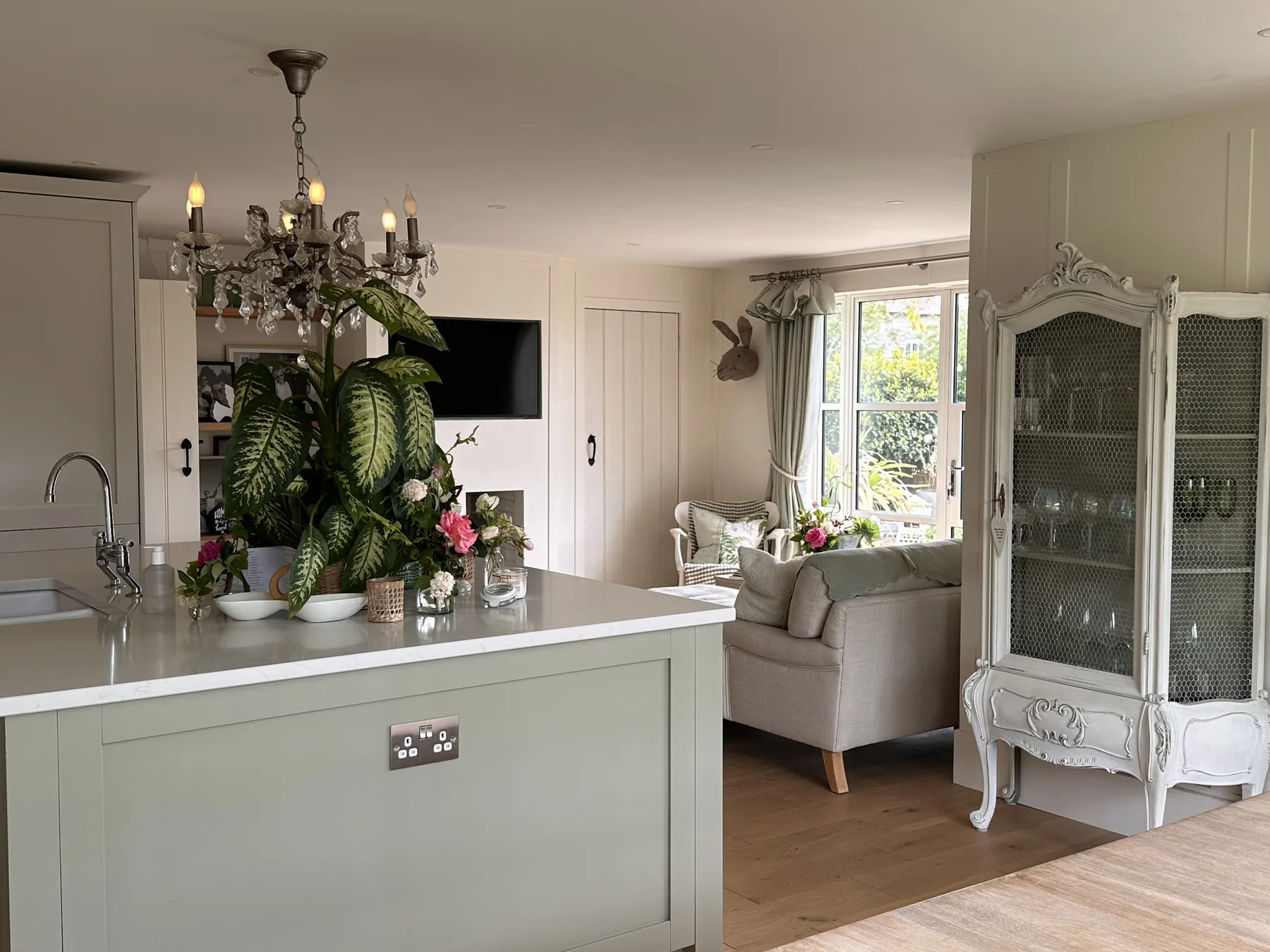 Open-plan kitchen and sitting room — sage cabinetry, French armoire, statement chandelier, and easy flow between living spaces
