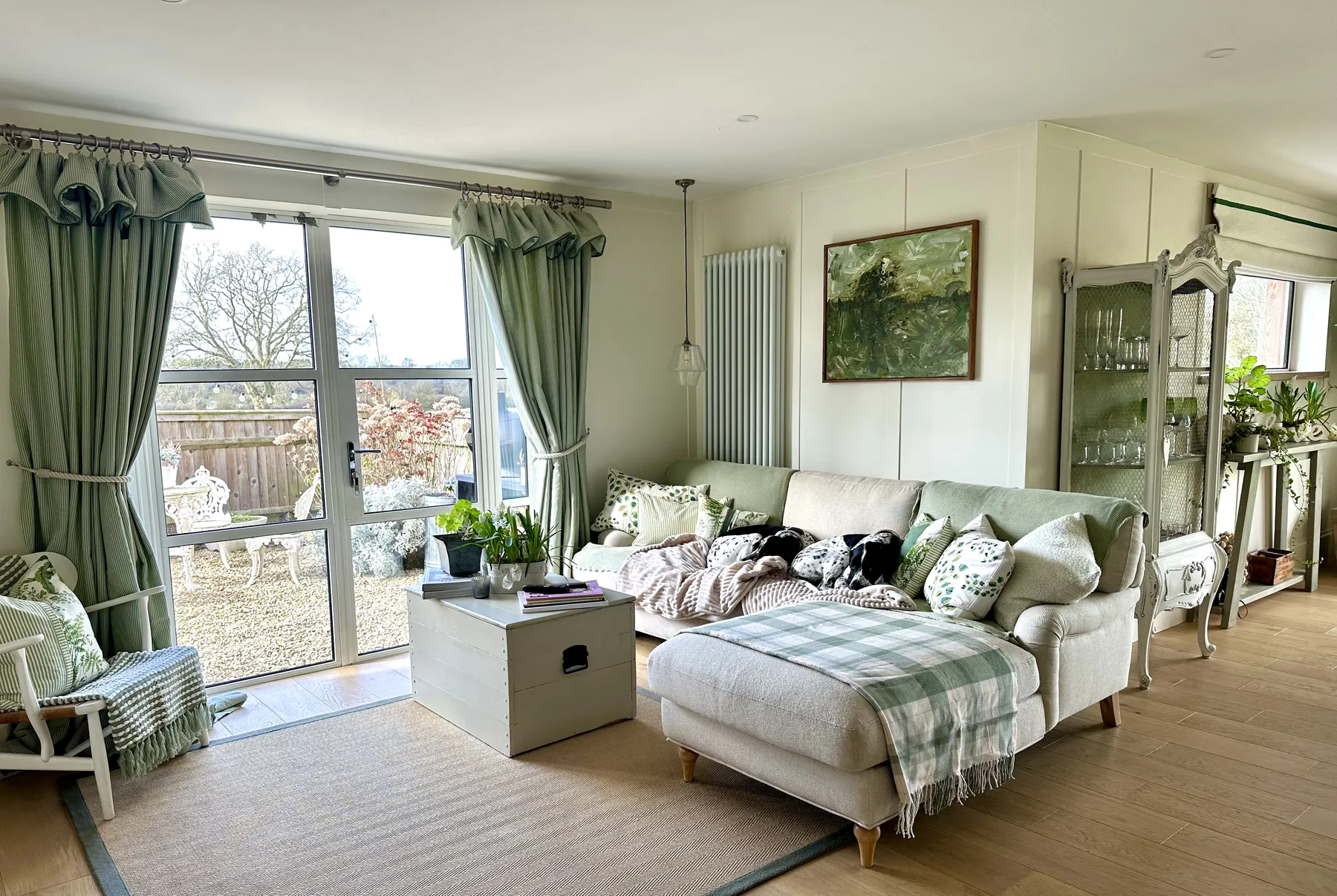 A second sitting room beyond the kitchen — soft linen sofa, layered checks and stripes, garden doors framed in green