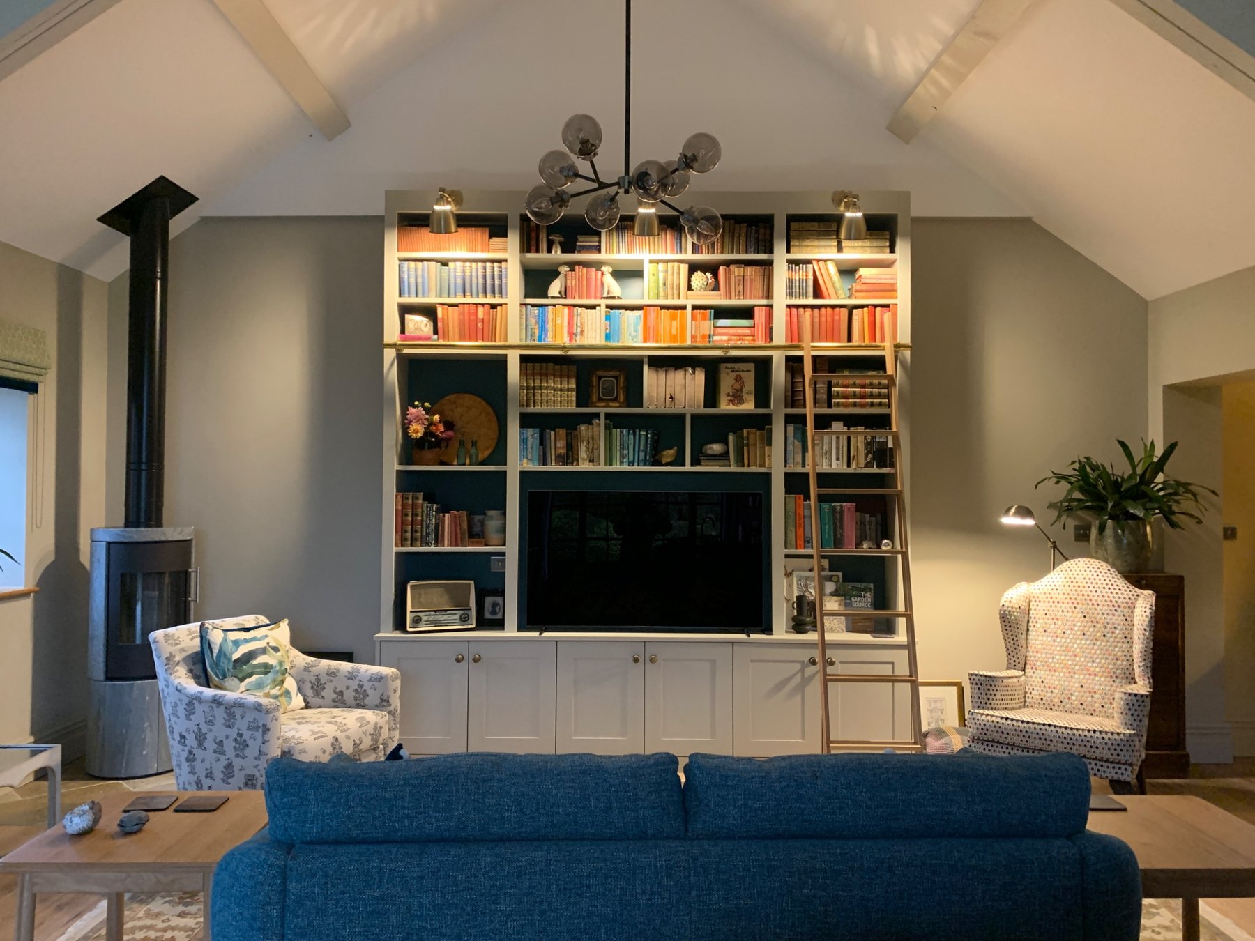 Full interior design project — vaulted living room with bespoke bookshelf wall, blue sofa, log burner and pitched beams