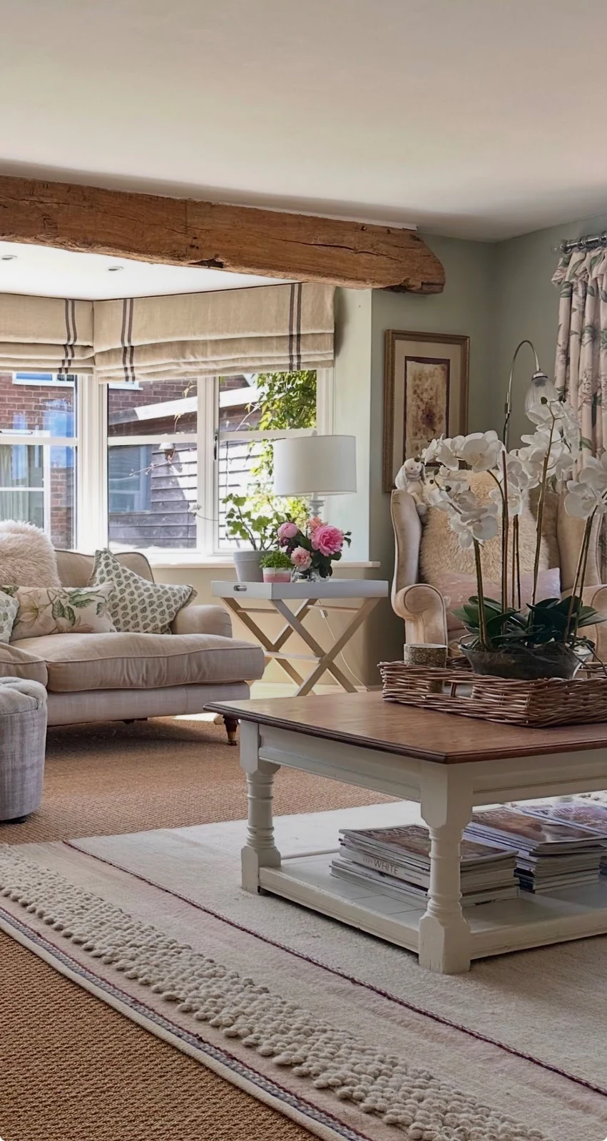 A beamed sitting room layered with linen blinds, a lamp, orchids and fresh flowers — the finishing details that make a furnished room feel finished